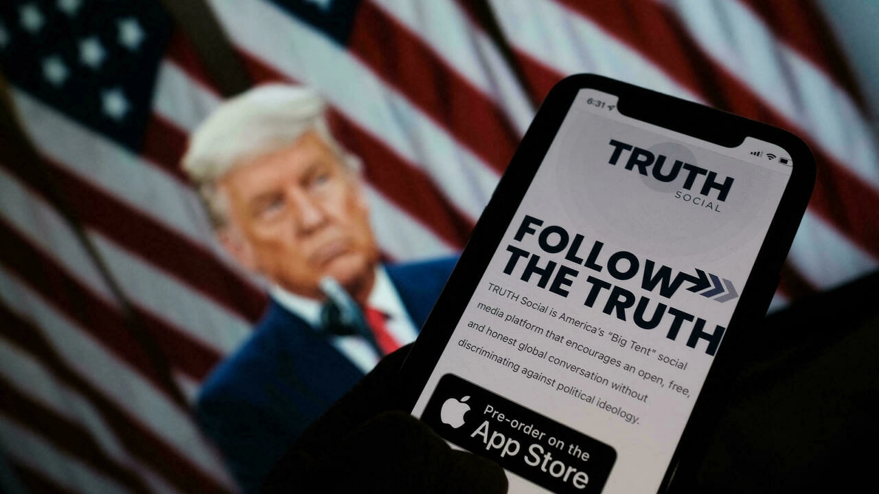 A person checking the application store on a smartphone for Truth Social, with a photo of former US president Donald Trump on a computer screen in the background, in Los Angeles, California, on October 20, 2021. © Chris Delmas, AFP (file photo)